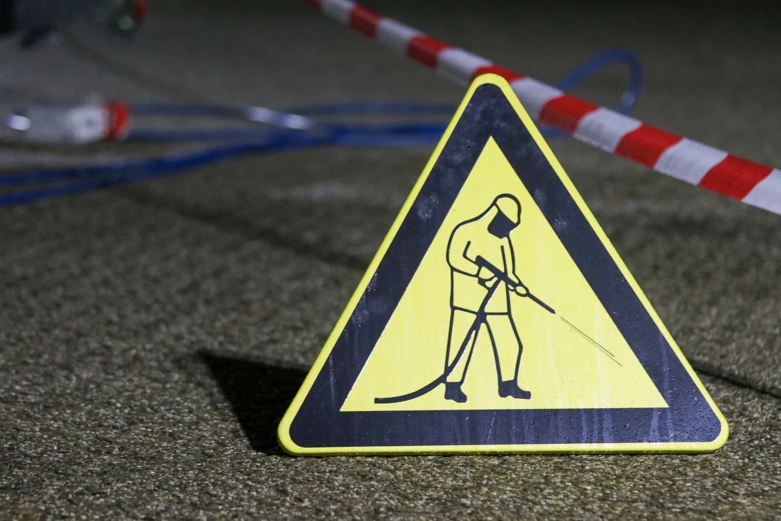 Yellow caution sign depicting pressure washing in a night setting with safety barriers. Signing off pressure washing staff: what the assessor watches for