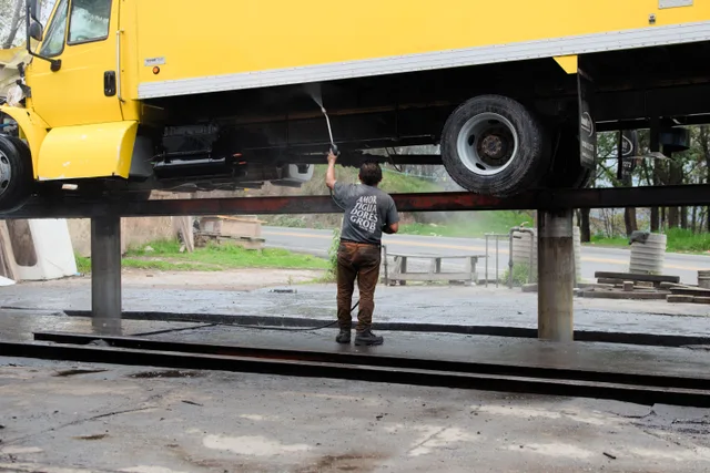 A worker power washes the underside of a raised yellow truck at an outdoor service station. Pressure washing fleet routing software: what to test first