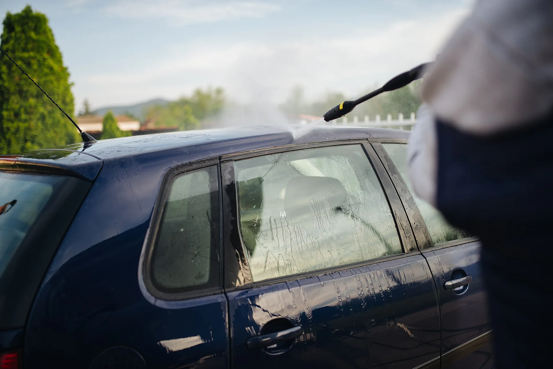 A person is washing a blue car in a residential driveway. Water spray from a pressure washer creates a mist under the warm sun, highlighting a clean and vibrant setting. Pressure washing KPIs: five pairs to read together each month