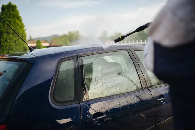 A person is washing a blue car in a residential driveway. Water spray from a pressure washer creates a mist under the warm sun, highlighting a clean and vibrant setting. Pressure washing KPIs: five pairs to read together each month